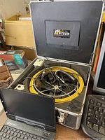 Photo showing Anysun inspection camera system inside a metal case with yellow cable reel and attached cables, placed on a table with miscellaneous items around it.