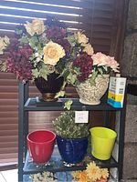 Wide view showing two large decorative vases with faux flowers and four empty or filled pots on a black shelf by window with wooden blinds.