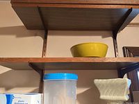 Wooden shelves with yellow mixing bowl, clear plastic storage container with blue lid, white ceramic planter, and plastic dinnerware set box