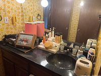 Wide bathroom countertop view showing various personal care items including perfume bottles, jewelry box, plug-in lights, hairdryer and assorted bottles next to the sink.