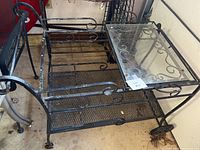 Full view of the black metal rolling cart showing glass top, expanded metal bottom shelf, decorative scrollwork, and wheels.