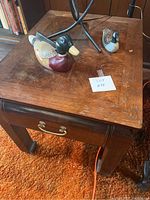 Photo showing wooden end table with two carved wooden duck figurines on top, visible wood grain and drawer.