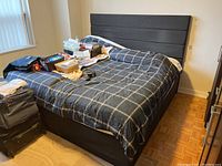 Full view of the king size bed frame with dark padded upholstery, showing bed skirt and mattress covered in a checked blanket with various items on bed.