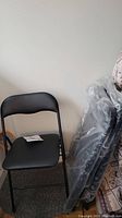One black vinyl folding chair out of packaging with 3 others still in clear plastic packaging leaning against wall.