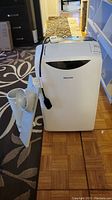 Front view of portable AC unit with attached power cord and nearby exhaust hose and vent panel