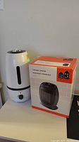 Photo showing the ceramic heater external unit next to its original packaging box, displaying the heater's compact size and design.