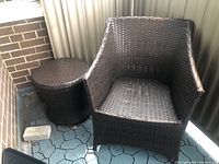 Outdoor plastic woven chair with cushion and matching woven table on balcony or patio with tiled floor and brick wall.