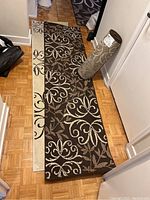 Two hall runners laid flat on a hardwood floor, one dark brown and one cream base, both with floral scroll designs. One rolled runner standing nearby.