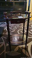 Full view of the oval wooden side table showing turned legs, bottom shelf, drawer with knob and decorative top supports.