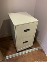 Front and side angle view of the small beige 2-drawer metal filing cabinet on rollers. Visible wear and some damage to edges and surface.