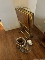Photo showing four stacked retro TV trays with wood-grain tops and brass frames, placed against a wall. On the floor in front of the trays are a white ceramic teapot, two planters (one ceramic and one glass), and a small decorative plaque.