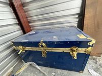 Blue metal steamer trunk closed, showing top and front corner with brass hardware and scratches.
