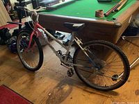 Full side view of the ladies bicycle showing pink and white frame, tires, pedals, seat, and part of the room interior including a pool table.