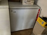 Front and side view of white Danby Premiere deep freezer next to another appliance and storage boxes on top.