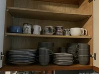 View inside wooden cabinet showing two shelves with stacked gray dishes and various mugs on top shelf.