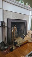 Photo showing two sets of fireplace tools and two metal firewood holders in front of fireplace.