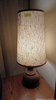 Photo of one lamp showing full height with cream-colored tall cylindrical fabric shade and wood surface
