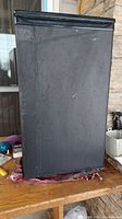Black mini fridge with a single door and weathered exterior placed on a wooden surface outside a house.