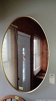 Front view of the oval mirror reflecting part of a door and window blinds, showing the metallic frame and clean mirror surface.