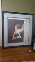 Framed print leaning against a green wall on wooden floor, showing full frame and mat with visible sticker on the top left corner.