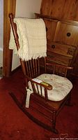Wooden rocking chair with white floral-patterned seat cushion and cream-colored knitted afghan draped over the backrest, set on a red carpet.