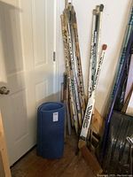Photo showing approximately 13 hockey sticks leaning against wall with two rolled blue yoga mats in front.