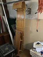 Two wooden toboggans leaning against a wall in a storage area, showing the full height and structure of the sleds including pull ropes and curved front of the larger one.