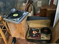 Overview of both record players: the Kelton Ultrasonics phonograph in a wooden suitcase with a record on its platter and external cord, next to the Taya MP-200 turntable on a wooden stand playing a record with a yellow label.