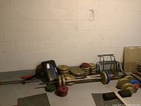 Photo showing a collection of multiple exercise weight plates of different sizes and colors stacked on the floor next to weightlifting bars and Curl Mate curling bar attachment.