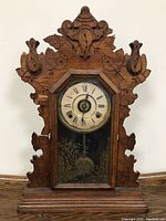 Full front view of antique wooden mantel clock showing carved wood details, glass door with Roman numeral clock face, and wood damage on edges.