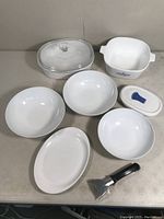 Lot overview showing white Corning Ware oval platter, three white bowls, blue cornflower casserole and lid, additional casserole with plastic lid, clear glass lid and utility handle.