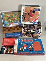 Group photo showing boxed board games and toy cars. Visible are Snoopy Game box, Lite-Brite box with child illustration, chessboard, and a colorful puzzle shaped as a US map. Below are toy cars in a blue container.