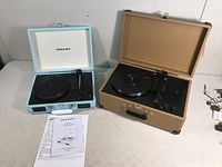 Two Crosley portable record players displayed open, showing turntables, tonearms, and control knobs with instruction sheet beside the blue model.
