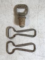 Three vintage Moxie soda bottle openers with varying wear; one is a bottle stopper with a metal opener top and cork base, the others are standard metal openers