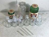 Photo showing eight clear glass bottles with ground stoppers including two larger labeled bottles and six smaller clear bottles, arranged on a surface with some label aging and one chipped bottle.