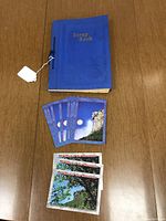Blue scrapbook book shown closed with several contemporary Old Man of The Mountain postcards laid next to it on a wooden surface.