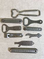 Nine vintage metal bottle openers laid out on a white surface, showing various shapes and embossed or engraved soda advertisements.