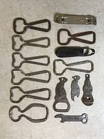 Top view of 14 bottle openers laid out in rows on white surface, showing varied metal openers including black handle one and ornate shaped ones.