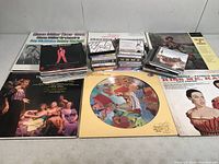 Collection showing vinyl albums, cassette tapes, and CDs stacked on table surface with visible album cover artwork including musicals and vintage record covers.