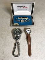 Three items shown: vintage Hamilton women's watch in original box with gold-tone bracelet, women's watch with moon phase dial and brown leather strap, metal quartz watch with black dial and attached compass and carabiner