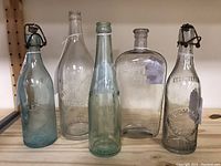 Five vintage and antique bottles on wooden shelf including clear and aqua glass with embossed lettering