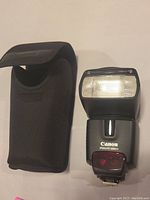 Canon Speedlite flash unit next to its carrying case, showing front view of flash head and case material and design.