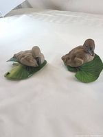 Front view of two brown China duck figurines positioned on green leaf bases, showing detailed feather patterns and lifelike postures.