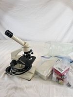Beige compound microscope angled to show eyepiece, objective lenses, and base with accompanying boxes of prepared slides and small plastic bags of accessories on white backdrop.