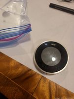 Photo showing the Nest Thermostat 2nd Generation on a white surface with part of a wooden table and a plastic bag nearby.