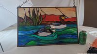 Stained glass panel with ducks, reeds, and sunset sky, hung by chain, shown with soda can for size reference.