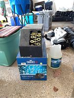 Black Chauvet DJ bubble machine sitting on its original box next to a jug of bubble fluid on a concrete floor, surrounded by random items.