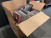 Box filled with approximately 40-50 music CDs standing upright inside a cardboard box, showing various jewel case spines with artists and titles.