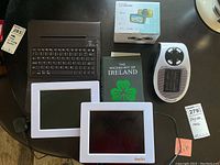 Overview showing both Dayclox digital calendar clocks, black keyboard case, boxed car dashcam, white wall plug space heater, and 'The Wicked Wit of Ireland' book laid on table.
