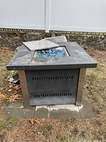 Full view of square propane firepit sitting on outdoor pavers with metal cover placed partially off, showing holds and rust spots on top surface.